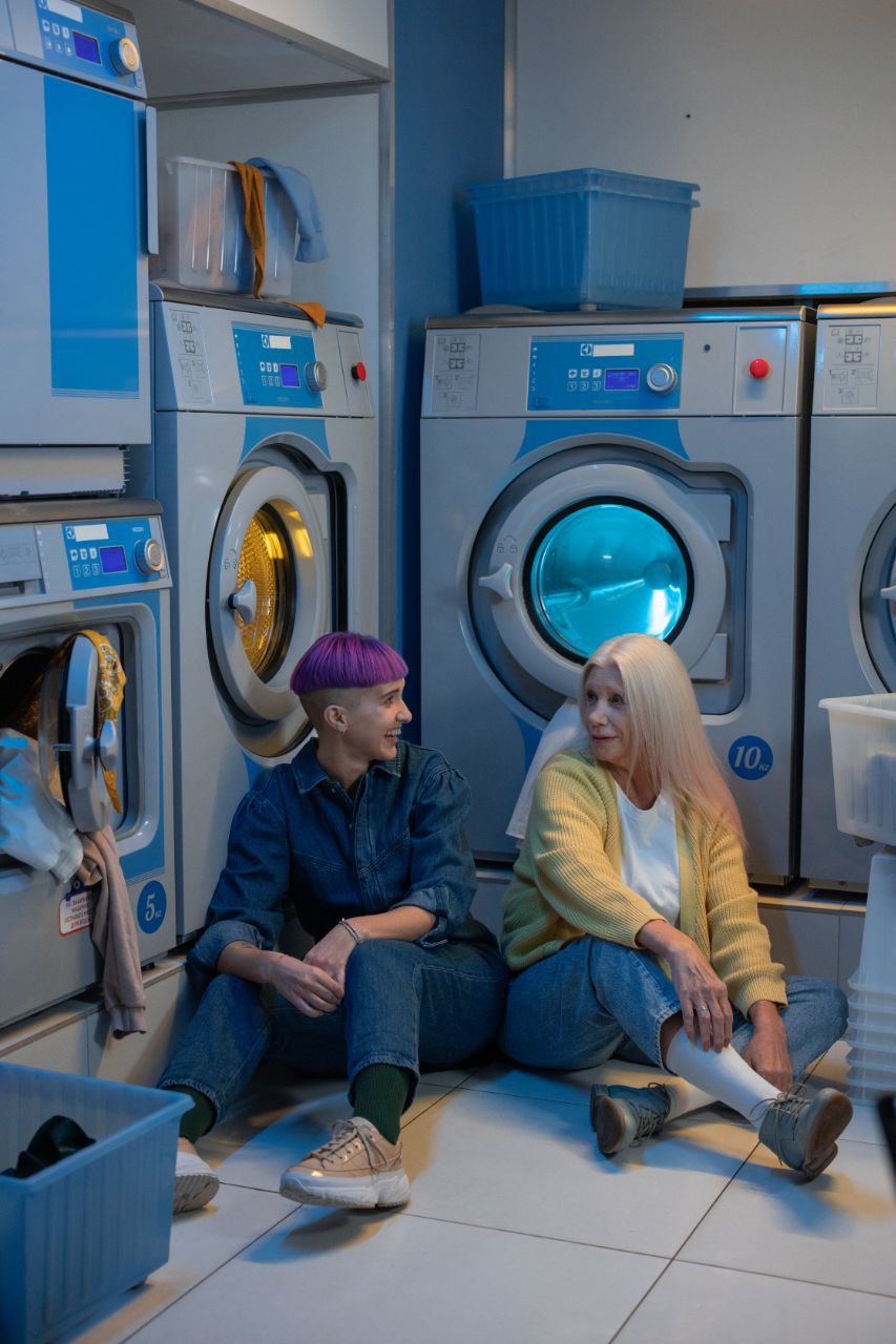 Two women sitting on the floor of a colourful laundromat, laughing together